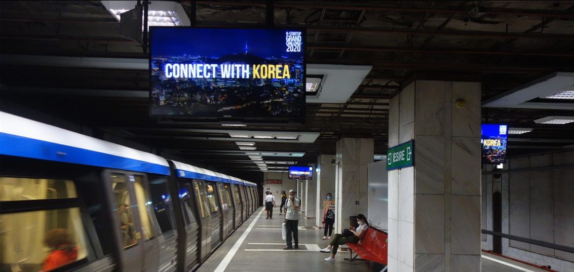 K-Startup Grand Challenge 2020 'Connect with Korea' creative on a subway platform screen in Bucharest, with masked commuters and an arriving train