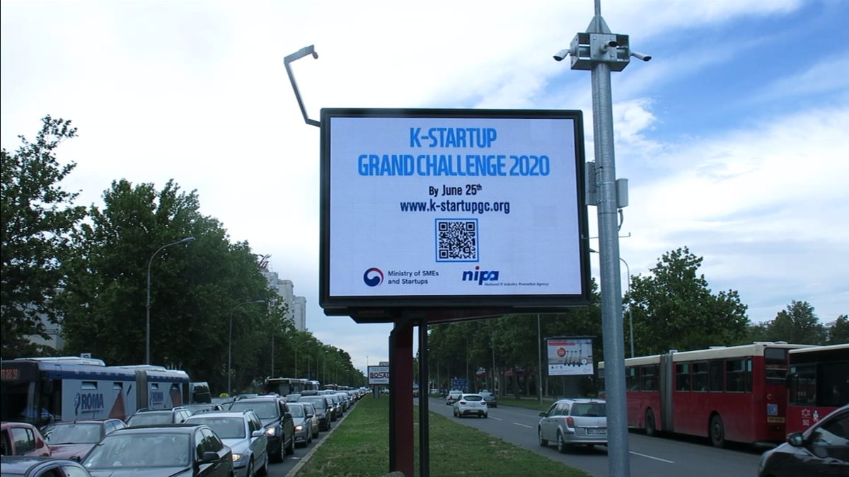 K-Startup Grand Challenge 2020 creative with the application deadline and QR code on a roadside digital billboard in Serbia, above heavy traffic