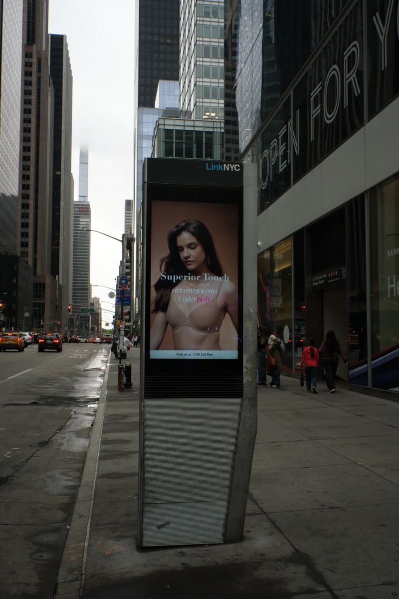 LinkNYC digital kiosk displaying an Intimissimi lingerie campaign with a store-visit call to action on a Midtown Manhattan sidewalk with yellow cabs and pedestrians