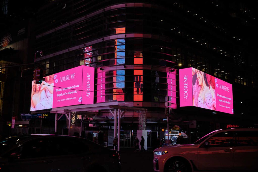 A digital billboard for Adore Me, illuminated in bright pink and featuring a model in lingerie with promotional text, positioned on the side of a building in a nighttime cityscape
