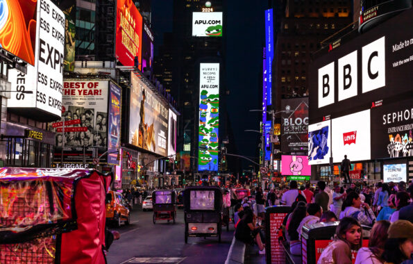 Crowded Times Square at night with glowing digital billboards covering both sides of the street above traffic and pedestrians. A tall central billboard stack shows "APU.COM" and cartoon frog graphics, while nearby signs for BBC, Levis, Sephora, and Broadway shows light the avenue.