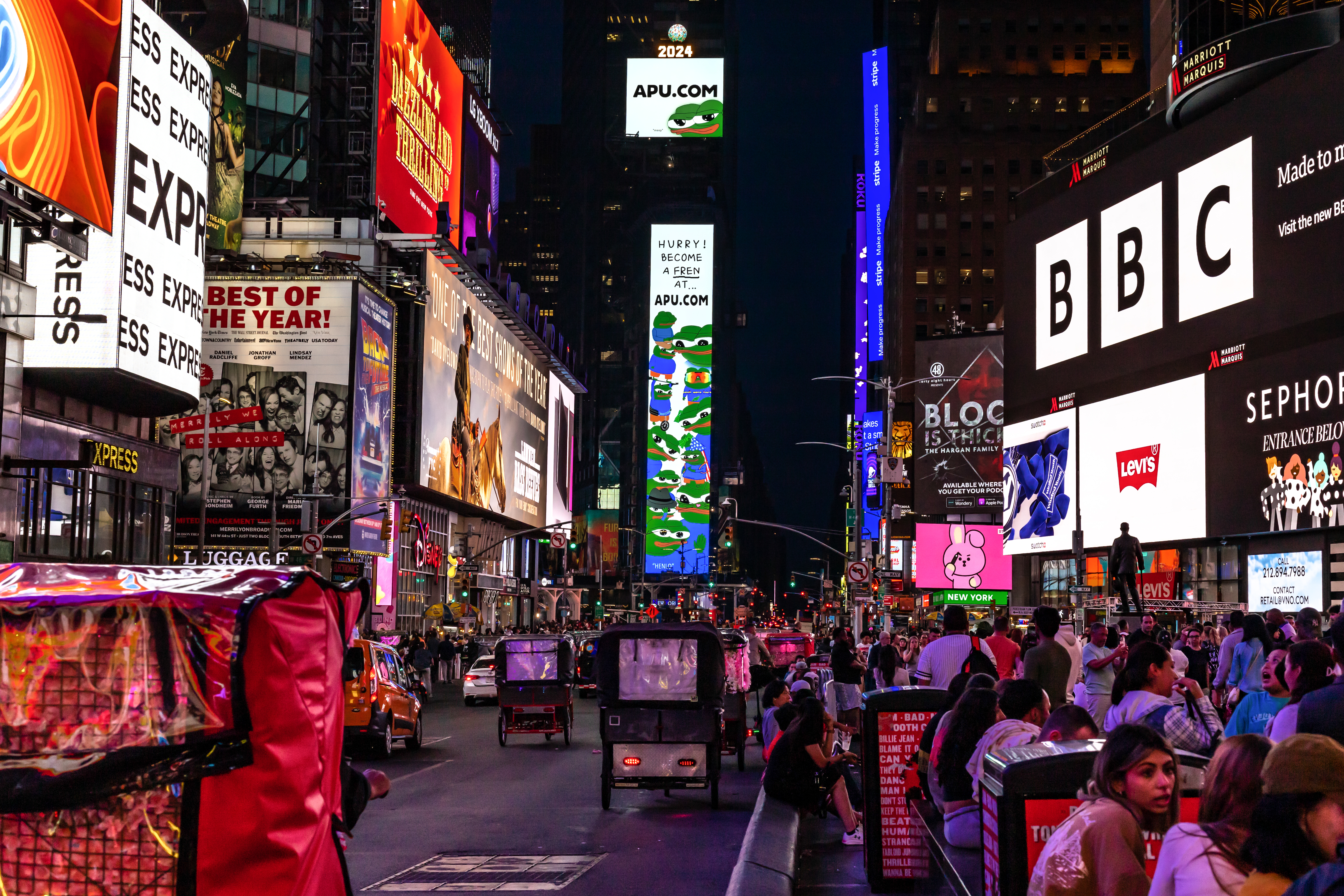 Crowded Times Square at night with glowing digital billboards covering both sides of the street above traffic and pedestrians. A tall central billboard stack shows "APU.COM" and cartoon frog graphics, while nearby signs for BBC, Levis, Sephora, and Broadway shows light the avenue.