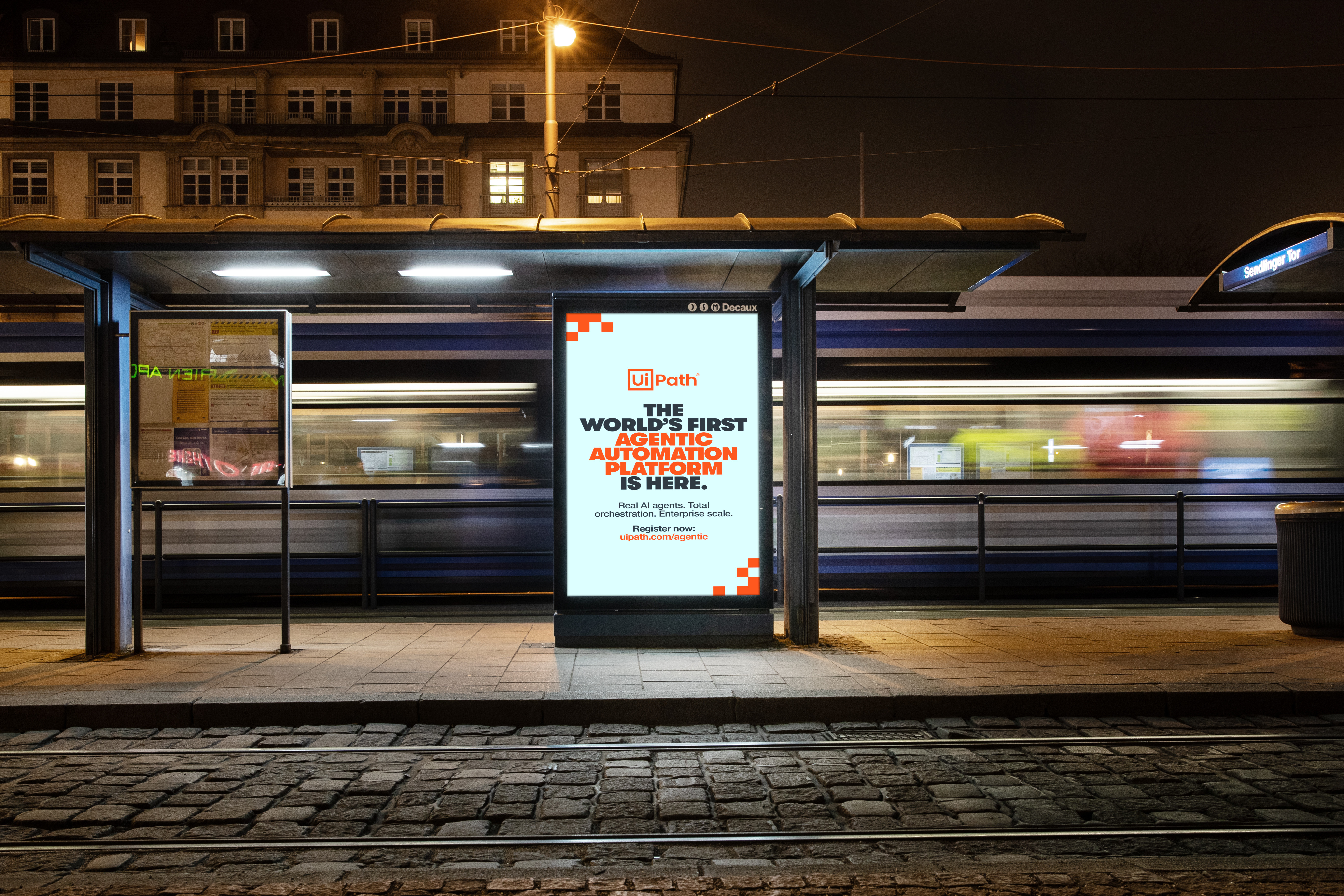 Nighttime tram stop digital billboard displays a UiPath ad that reads "THE WORLD'S FIRST AGENTIC AUTOMATION PLATFORM IS HERE." Smaller text below reads "Real AI agents. Total orchestration. Enterprise scale." and "Register now. uipath.com/agentic." A tram blurs past behind the shelter while warm streetlights and overhead wires frame the station.