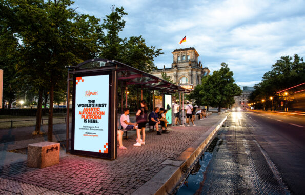 The 10-4 Shift: Why Growth Marketers are Ditching Agencies for DIY Rush Hour DOOH Bus shelter digital billboard beside a tree lined boulevard displays a UiPath ad that reads "THE WORLD'S FIRST AGENTIC AUTOMATION PLATFORM IS HERE." Several people sit and stand under the shelter while the Reichstag building rises in the background with the German flag above it and car lights streak along the wet street at dusk.