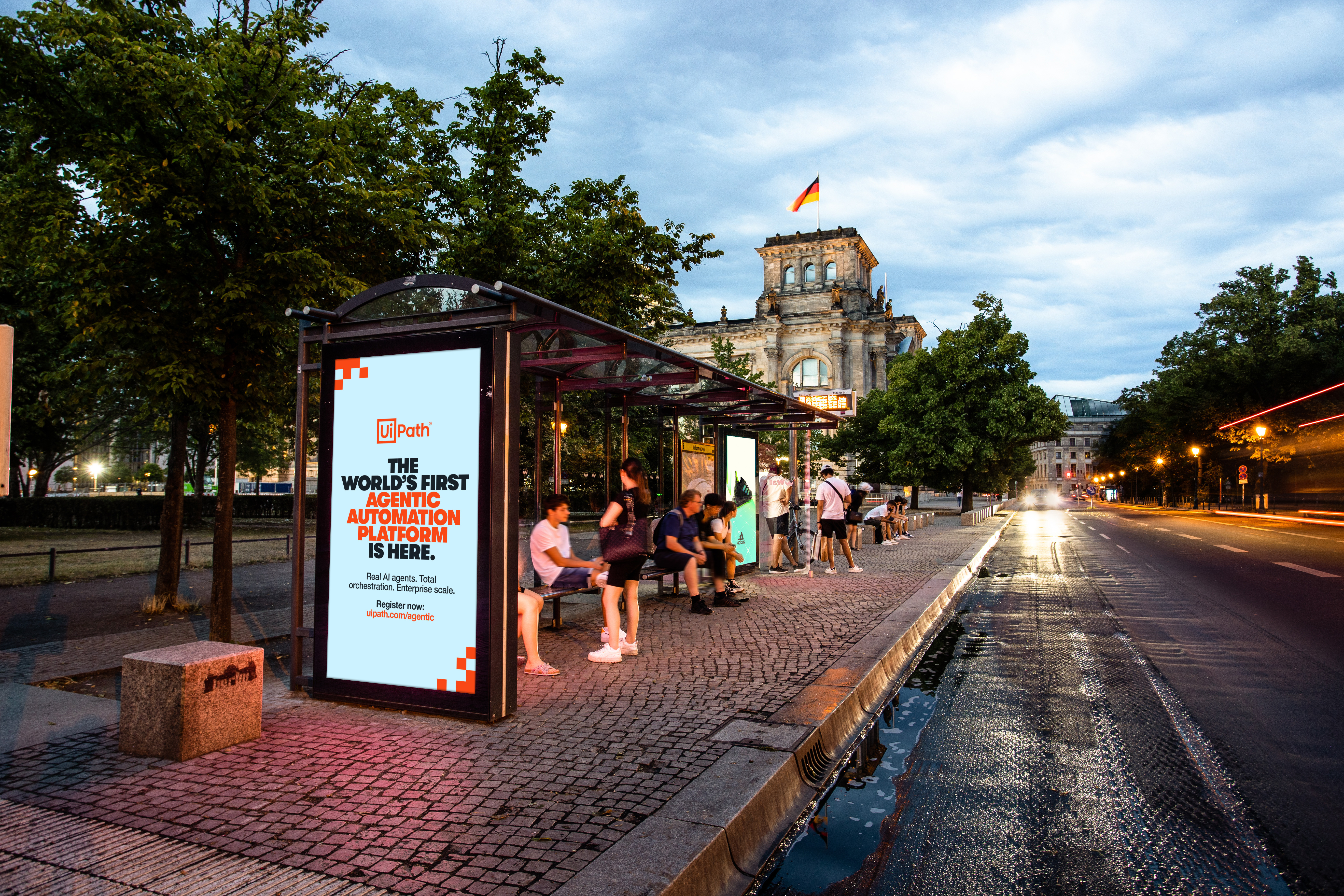 Bus shelter digital billboard beside a tree lined boulevard displays a UiPath ad that reads "THE WORLD'S FIRST AGENTIC AUTOMATION PLATFORM IS HERE." Several people sit and stand under the shelter while the Reichstag building rises in the background with the German flag above it and car lights streak along the wet street at dusk.