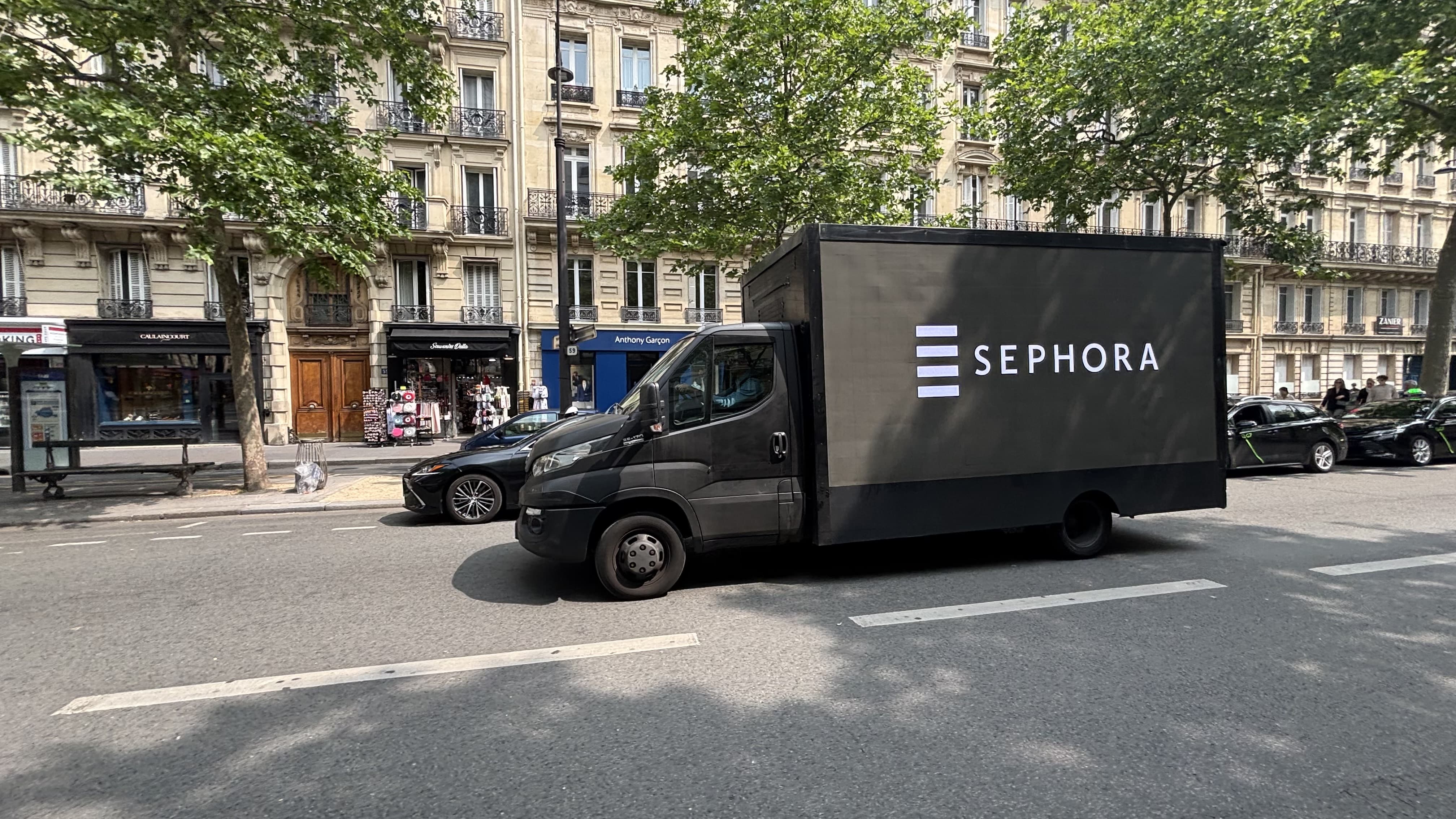 Black mobile billboard truck drives down a tree lined city boulevard with the white "SEPHORA" logo displayed across the side panel. Elegant Paris style apartment buildings, storefronts, and passing cars line the street behind it.