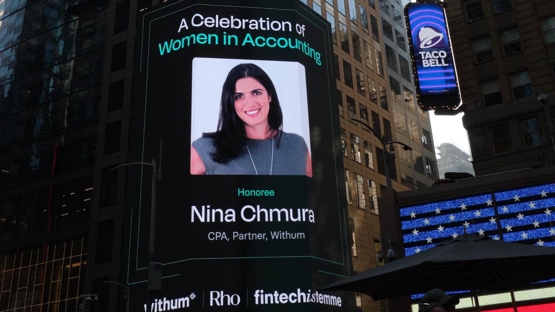 Times Square large-format digital billboard honoring Nina Chmura, CPA Partner at Withum, as part of the 'Celebration of Women in Accounting' campaign sponsored by Withum, Rho, and Fintechistemme, alongside Taco Bell signage and the American flag LED display in Midtown Manhattan, New York City.